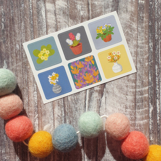 A set of six square envelope stickers with various spring flower designs displayed on top of a wooden surface surrounded by colorful pom-poms.