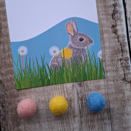 gift note sheet with a rabbit illustration on grass and dandelions, with three decorative felt balls below on a wooden surface.
